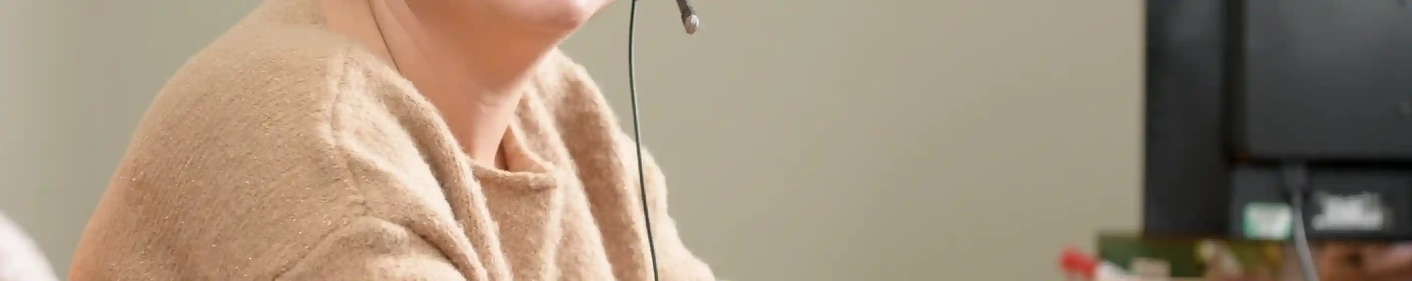 A woman with short brown hair and glasses sits at a desk wearing a headset, smiling at the camera. In the background, a banner for "Shorefield Holidays" is visible along with a computer and a decorative mug.