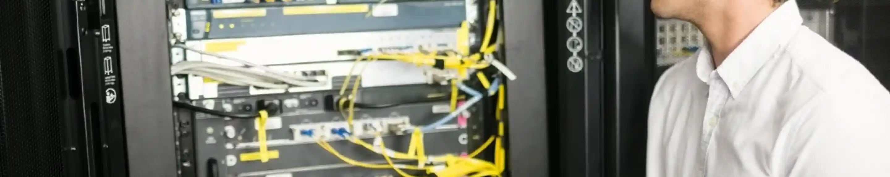 A technician in a white shirt operates a laptop while examining a server rack filled with network cables and equipment.