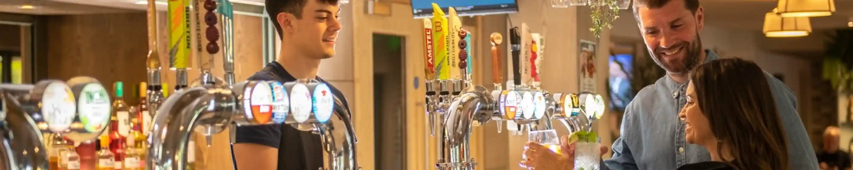 A bartender serves a drink to a woman at a modern bar while a man stands nearby smiling. The bar features several taps and hanging glassware, with a cozy and inviting atmosphere.
