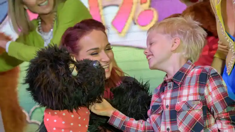 A young boy in a plaid shirt interacts joyfully with a woman holding a black puppet. They both smile and appear to be engaged in a playful moment, with colourful animated backgrounds behind them.
