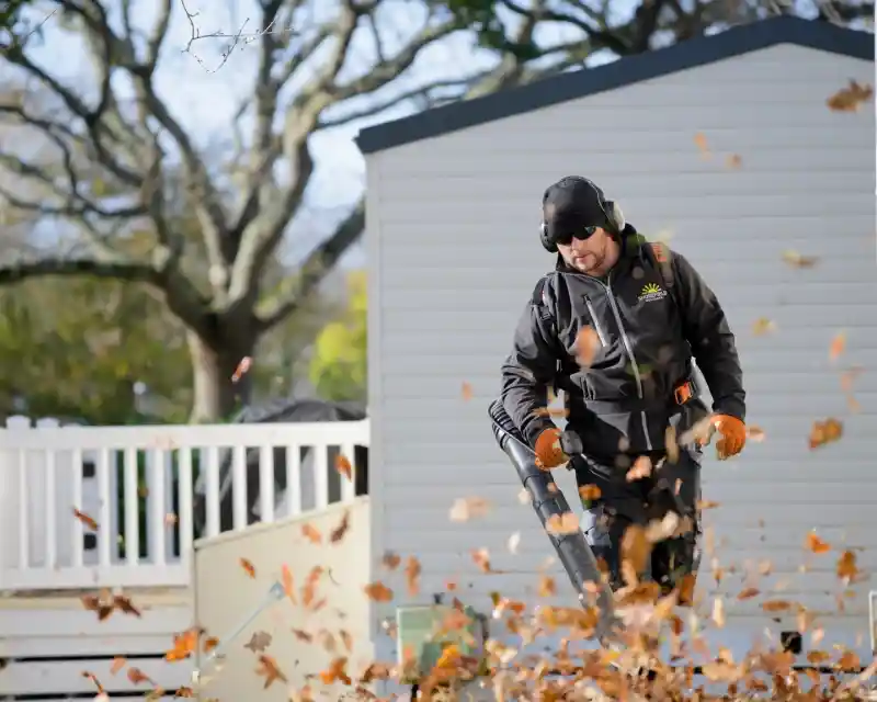 A person wearing a black jacket and headphones operates a leaf blower, sending leaves flying in the air. In the background, a shed and trees are visible, with sunlight illuminating the scene.