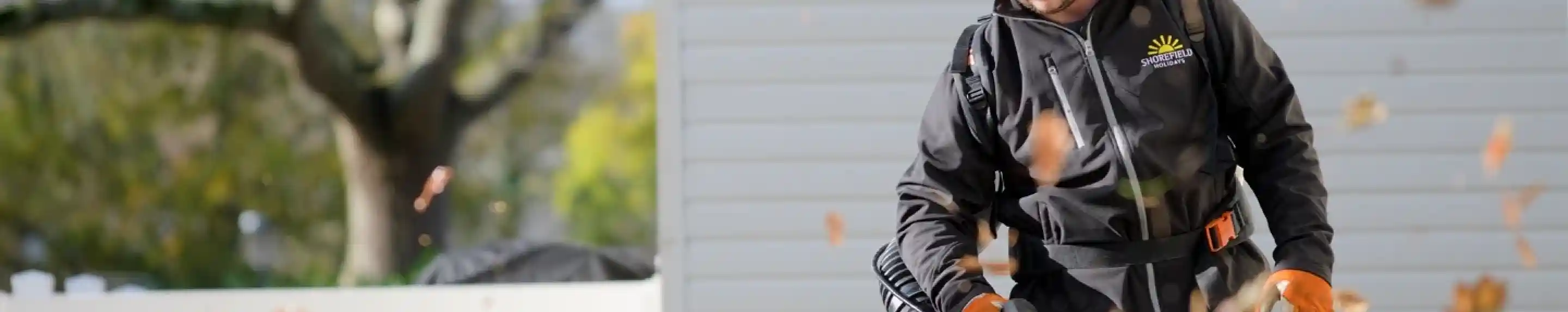 A person wearing a black jacket and headphones operates a leaf blower, sending leaves flying in the air. In the background, a shed and trees are visible, with sunlight illuminating the scene.
