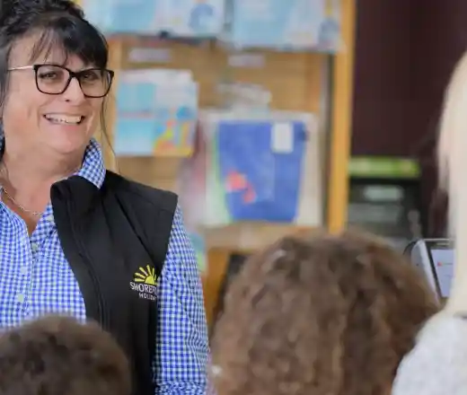A smiling woman wearing glasses and a black vest stands in front of a group of children, engaging in conversation. The background features shelves with various colorful items.