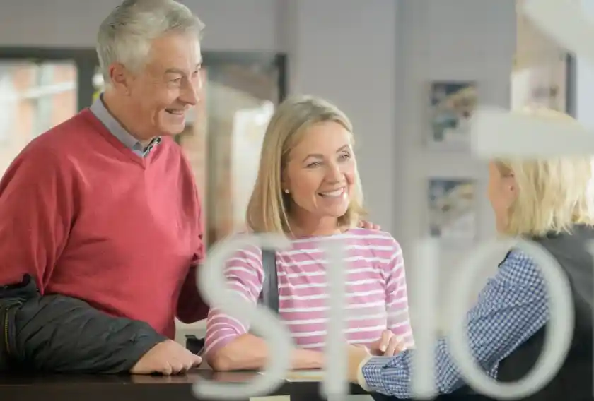 A smiling elderly couple interacts with a staff member at a reception desk. The man is wearing a red sweater, and the woman has on a striped shirt. They appear to be discussing something with the staff member, who is looking at them attentively.