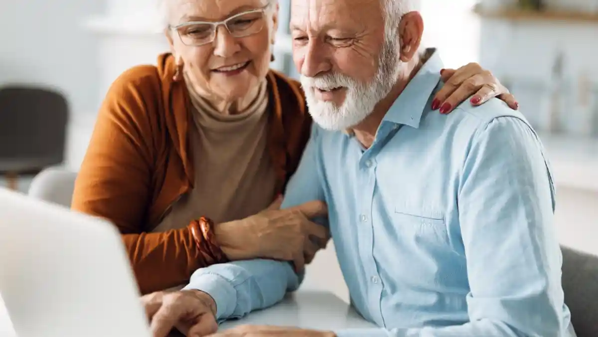 A smiling elderly couple interacts with a staff member at a reception desk. The man is wearing a red sweater, and the woman has on a striped shirt. They appear to be discussing something with the staff member, who is looking at them attentively.