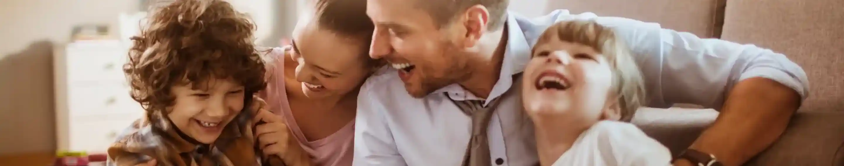 A father and three children are sitting on a carpet, laughing and playing together. The father holds a tablet as the kids enjoy the moment, sharing smiles and joy in a cosy living room setting.