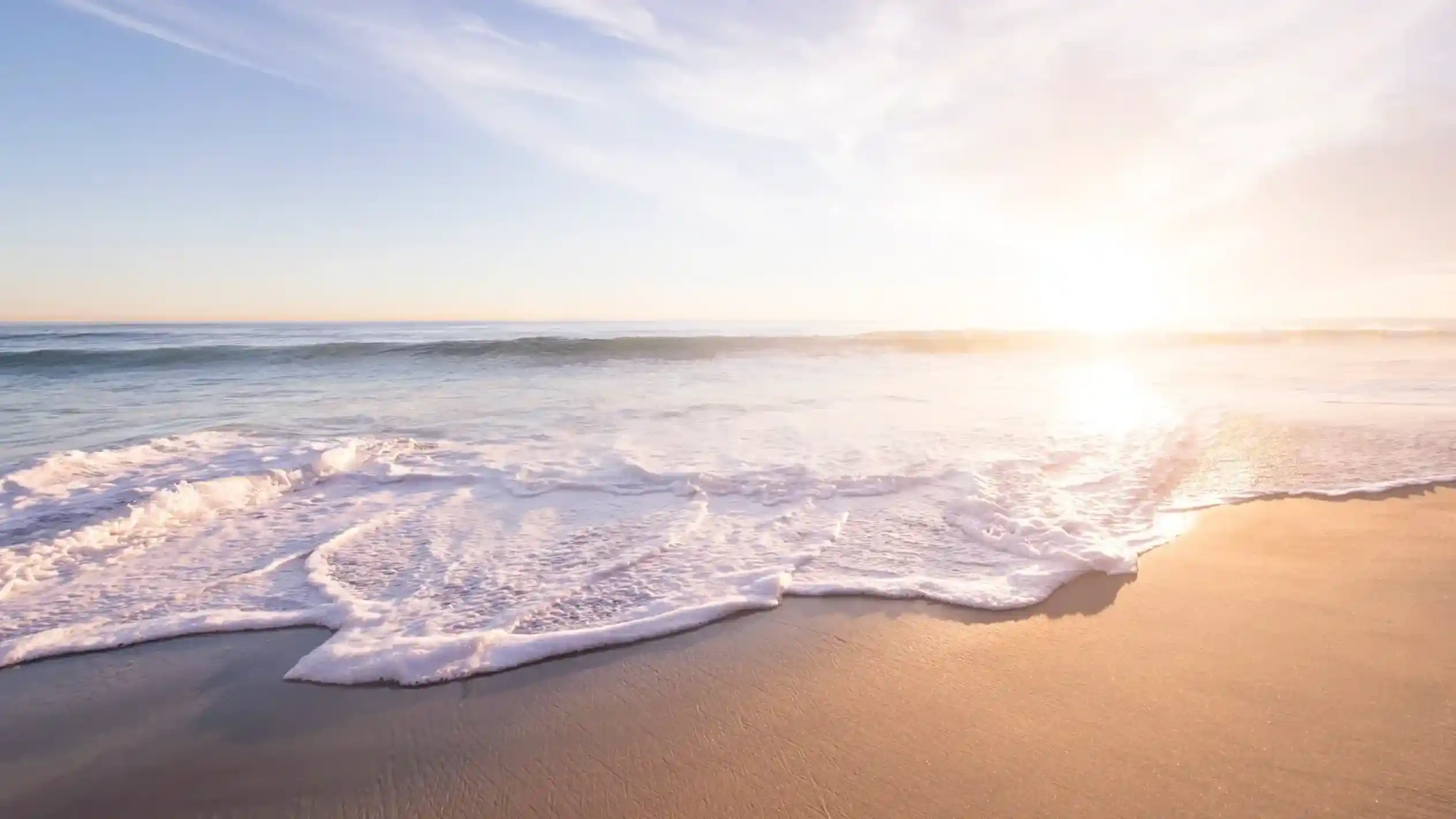 Gentle waves lapping at a sandy beach under a golden sunrise, with soft clouds in a clear sky.