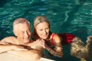 An older couple relaxes in a swimming pool, smiling and enjoying each other's company. The man has short gray hair and leans on the pool's edge, while the woman, in a red swimsuit, floats nearby. Sunlight reflects off the water, creating a serene atmosphere.