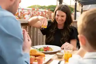 A woman sits at an outdoor dining table, smiling as she squeezes lemon over her meal. A plate with salad and colorful vegetables is in front of her. A young boy is seen in the foreground, and another person sits across from her, with a glass of drink on the table. The setting features greenery and a cozy atmosphere.