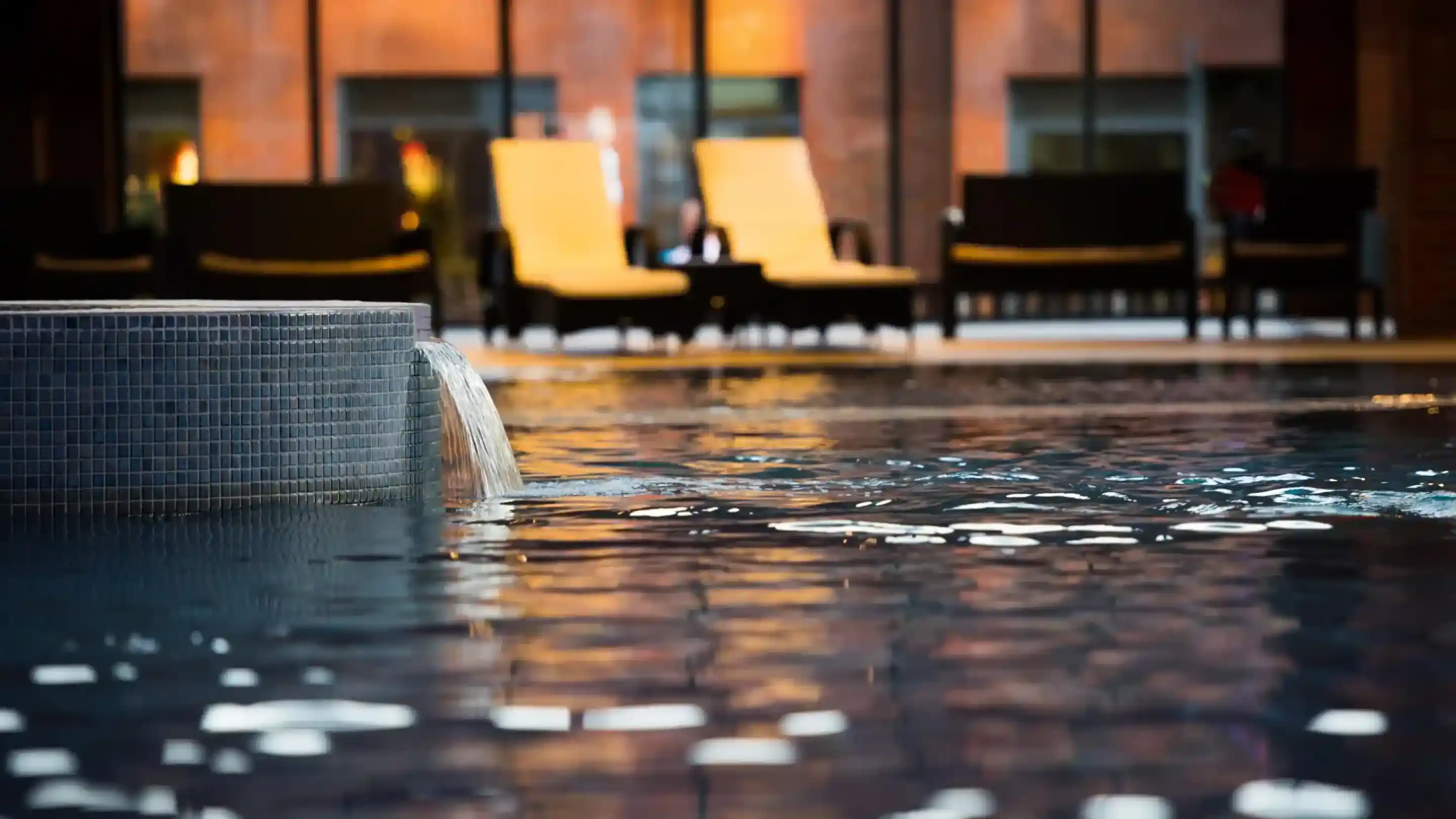 A tranquil indoor pool with a gentle water feature, surrounded by lounge chairs and large windows that reflect warm, ambient light.
