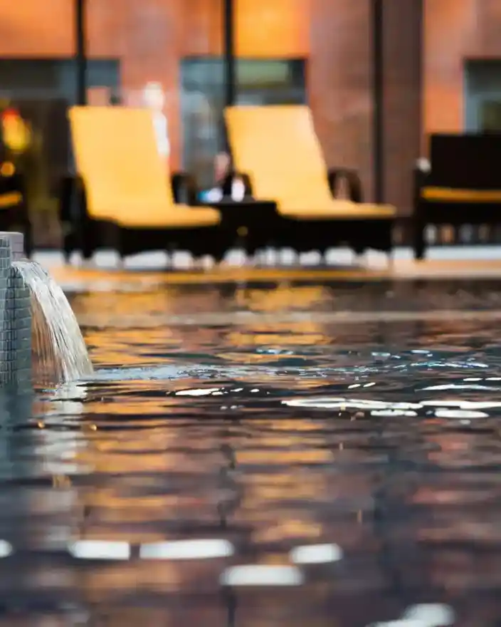 A tranquil indoor pool with a gentle water feature, surrounded by lounge chairs and large windows that reflect warm, ambient light.