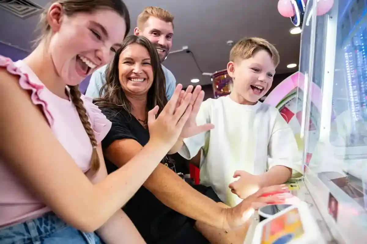 A family enjoys playing a carnival game together, smiling and laughing. Two children and two adults are engaged, with their hands on the game controls. Bright colors and arcade decorations are in the background.