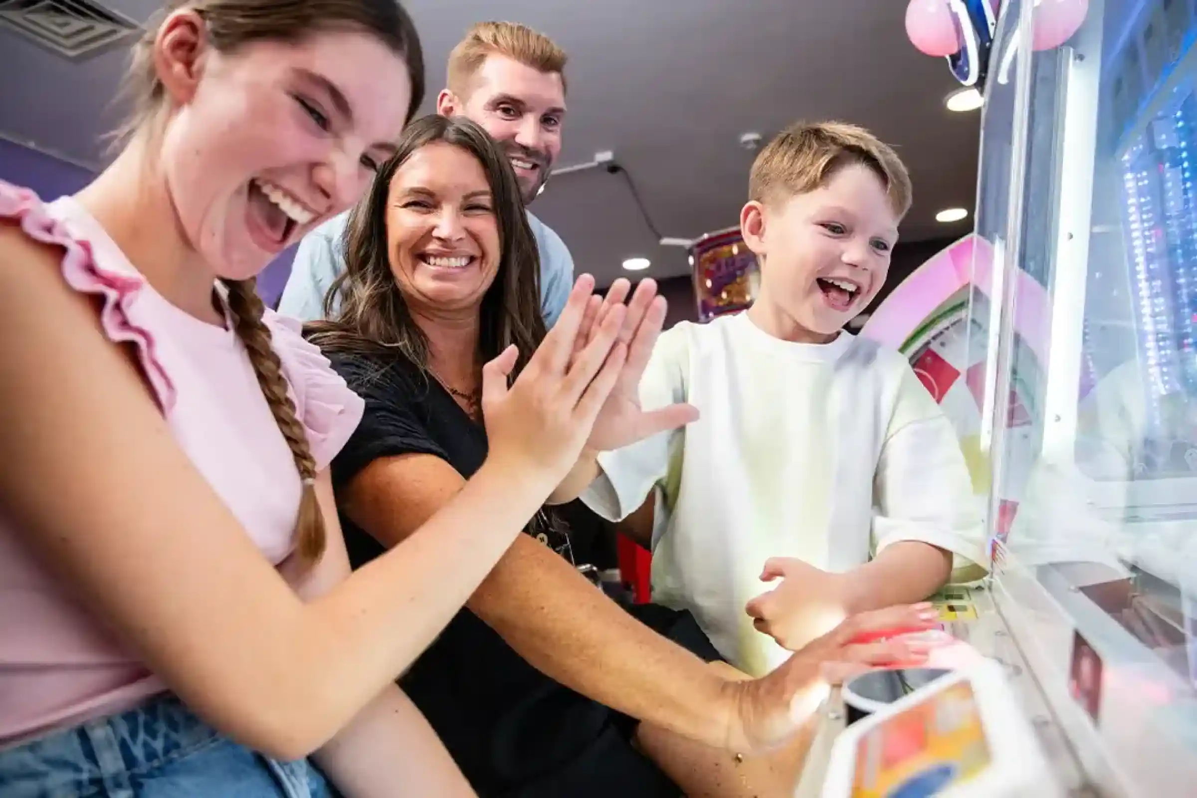 A family enjoys playing a carnival game together, smiling and laughing. Two children and two adults are engaged, with their hands on the game controls. Bright colors and arcade decorations are in the background.