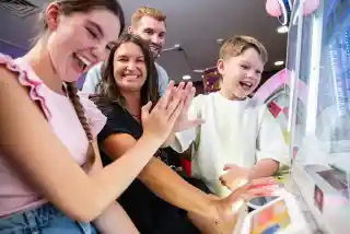 A family enjoys playing a carnival game together, smiling and laughing. Two children and two adults are engaged, with their hands on the game controls. Bright colors and arcade decorations are in the background.