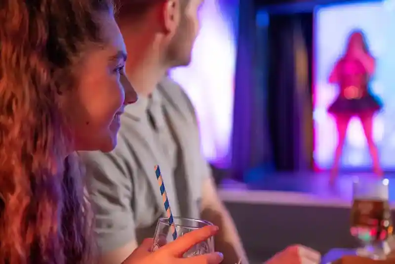A young woman with curly hair smiles while holding a drink with a striped straw. In the background, a person performs on stage under colorful lights. A man sits next to her, partially in view.