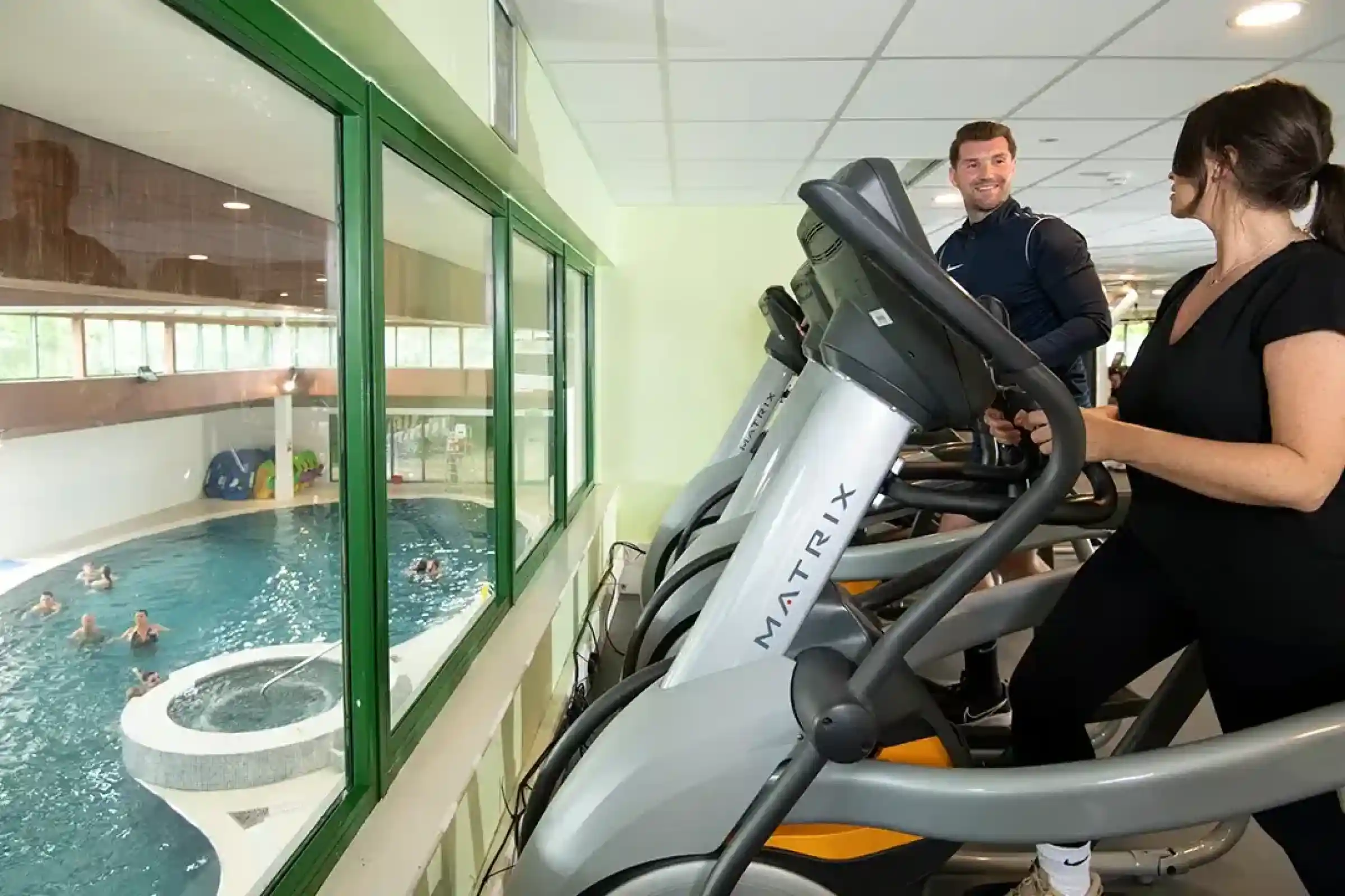 Two people exercising on elliptical machines in a gym with large windows overlooking an indoor swimming pool. One person is smiling while looking at the other. There are several individuals swimming in the pool.
