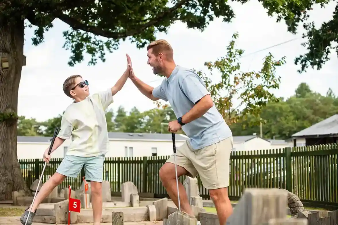 A child in sunglasses and a light shirt high-fives a smiling adult in a casual outfit while playing mini-golf outdoors. Green trees and a wooden fence are visible in the background.