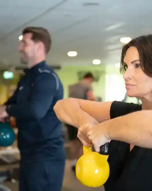 A woman and a man are exercising in a gym, both holding kettlebells. The woman is focused and performing a kettlebell squat, while the man is seen in the background, also engaged in a workout. The gym has various fitness equipment and a TV screen displaying content.