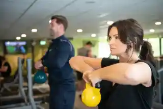 A woman and a man are exercising in a gym, both holding kettlebells. The woman is focused and performing a kettlebell squat, while the man is seen in the background, also engaged in a workout. The gym has various fitness equipment and a TV screen displaying content.