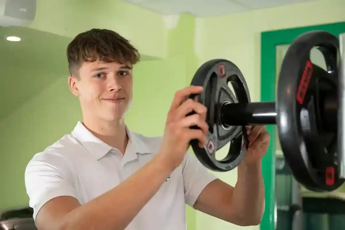 A young man in a white polo shirt adjusts weights on a barbell in a gym. He is smiling and appears focused on his task. The gym background features green walls and workout equipment.