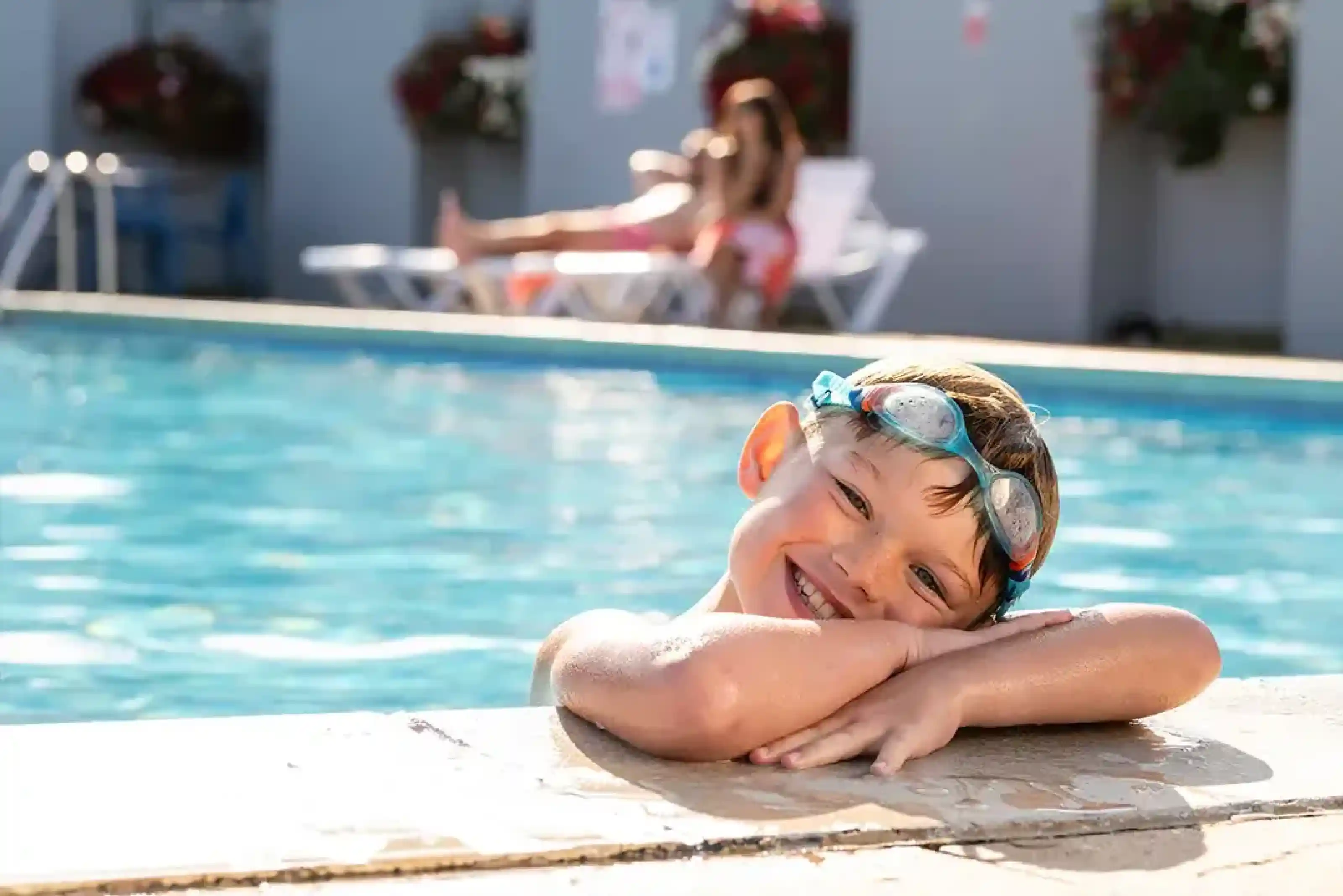 A smiling boy with swimming goggles leans over the edge of a swimming pool. In the background, a woman relaxes on a lounge chair. Bright sunlight reflects off the water.