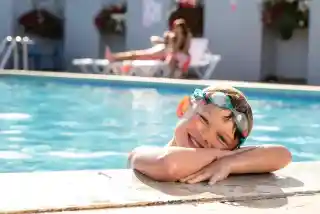 A smiling boy with swimming goggles leans over the edge of a swimming pool. In the background, a woman relaxes on a lounge chair. Bright sunlight reflects off the water.