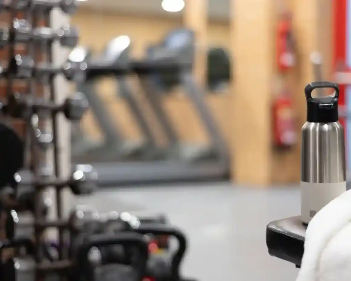 A stainless steel water bottle sits on a bench in a gym. In the background, there are treadmills and various weights on a rack. The space is well-lit and has a modern fitness ambiance.