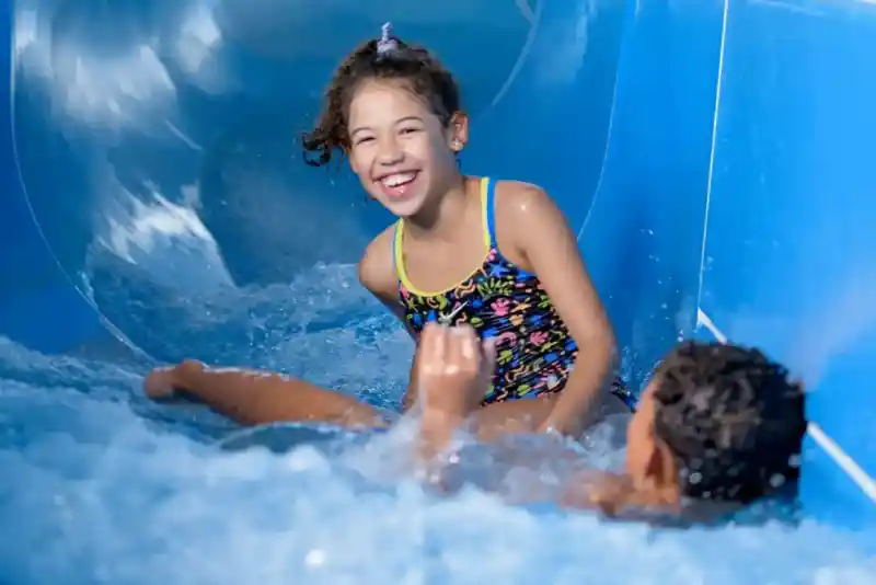 A young girl with curly hair enjoys a water slide, laughing and splashing water as she slides down. Another child is partially visible at the bottom of the slide, also in the water, creating a joyful atmosphere.