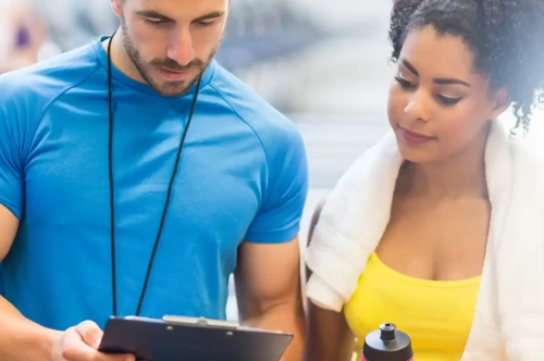 A fitness trainer and a female client discuss notes on a clipboard while standing in a gym. The trainer is wearing a blue shirt, and the client has a towel around her neck and a water bottle in her hand, wearing a yellow tank top.