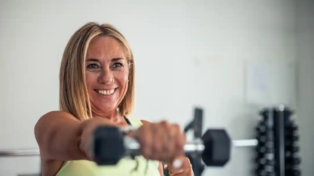 A smiling woman with shoulder-length blonde hair holds a dumbbell towards the camera, showcasing her strength and enthusiasm for fitness. The background features gym equipment, emphasizing a workout environment.