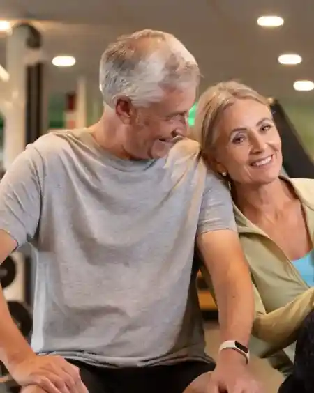 A smiling older couple sits together in a gym. The man, in a gray shirt, looks at the woman, who holds a green-top water bottle. They appear relaxed and happy.