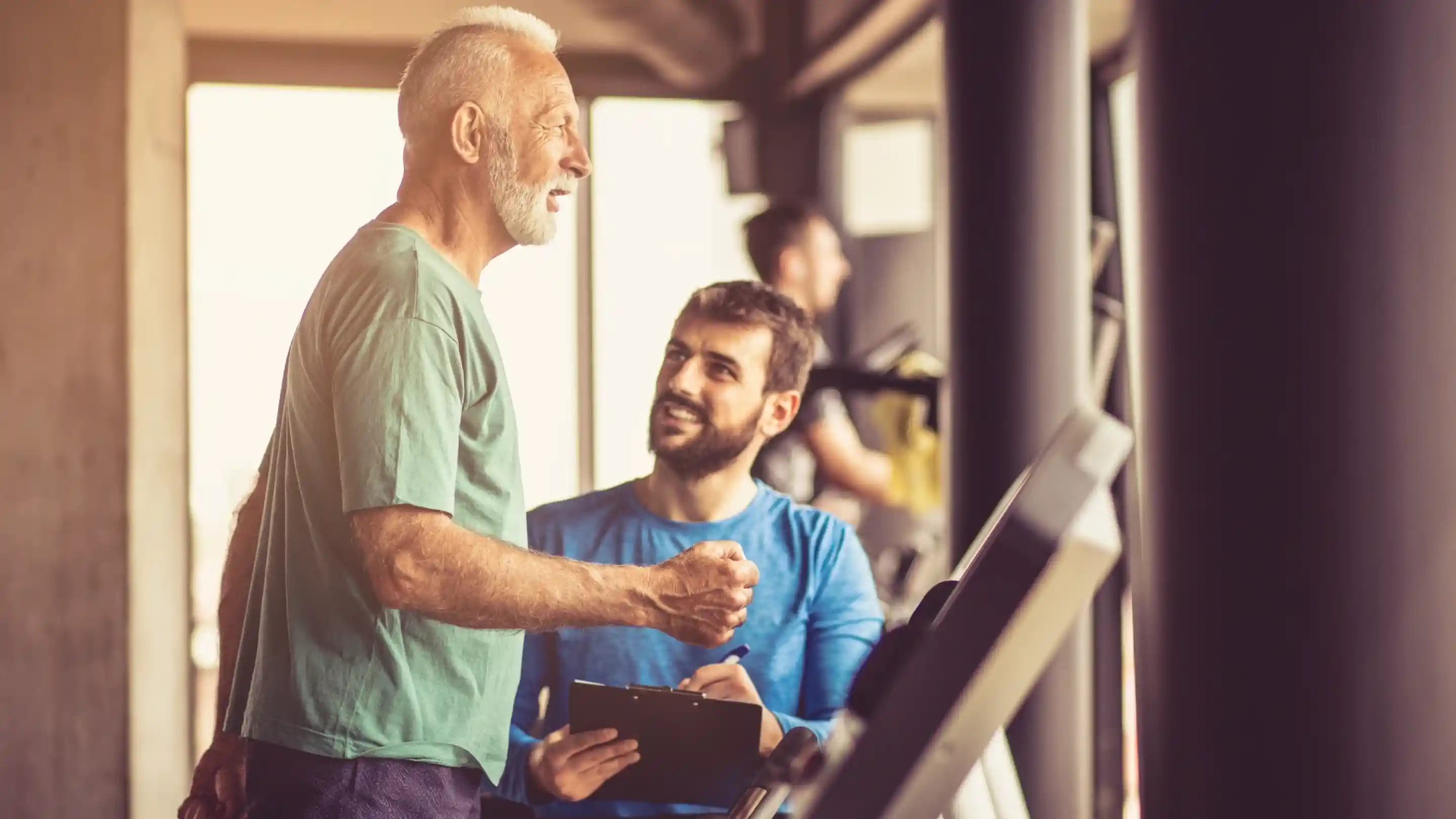 An older man in a green shirt smiles while talking to a younger man in a blue shirt, who is holding a clipboard. The scene is set in a gym with workout equipment in the background.