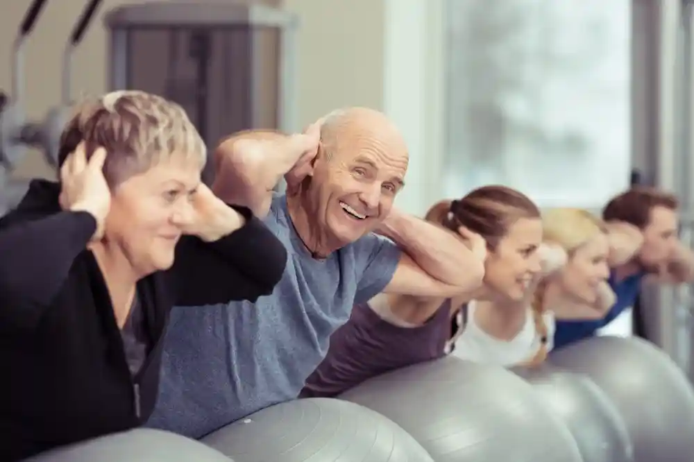 A group of five older adults, including a smiling elderly man, are exercising together on stability balls. They are engaging in a fitness class, holding their ears with their hands while leaning back. The atmosphere is cheerful and active, with gym equipment visible in the background.
