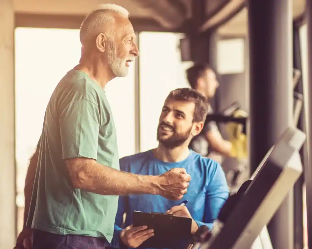 A senior man laughs and engages in conversation with a younger man at a gym. The younger man holds a clipboard and smiles, while others exercise in the background.