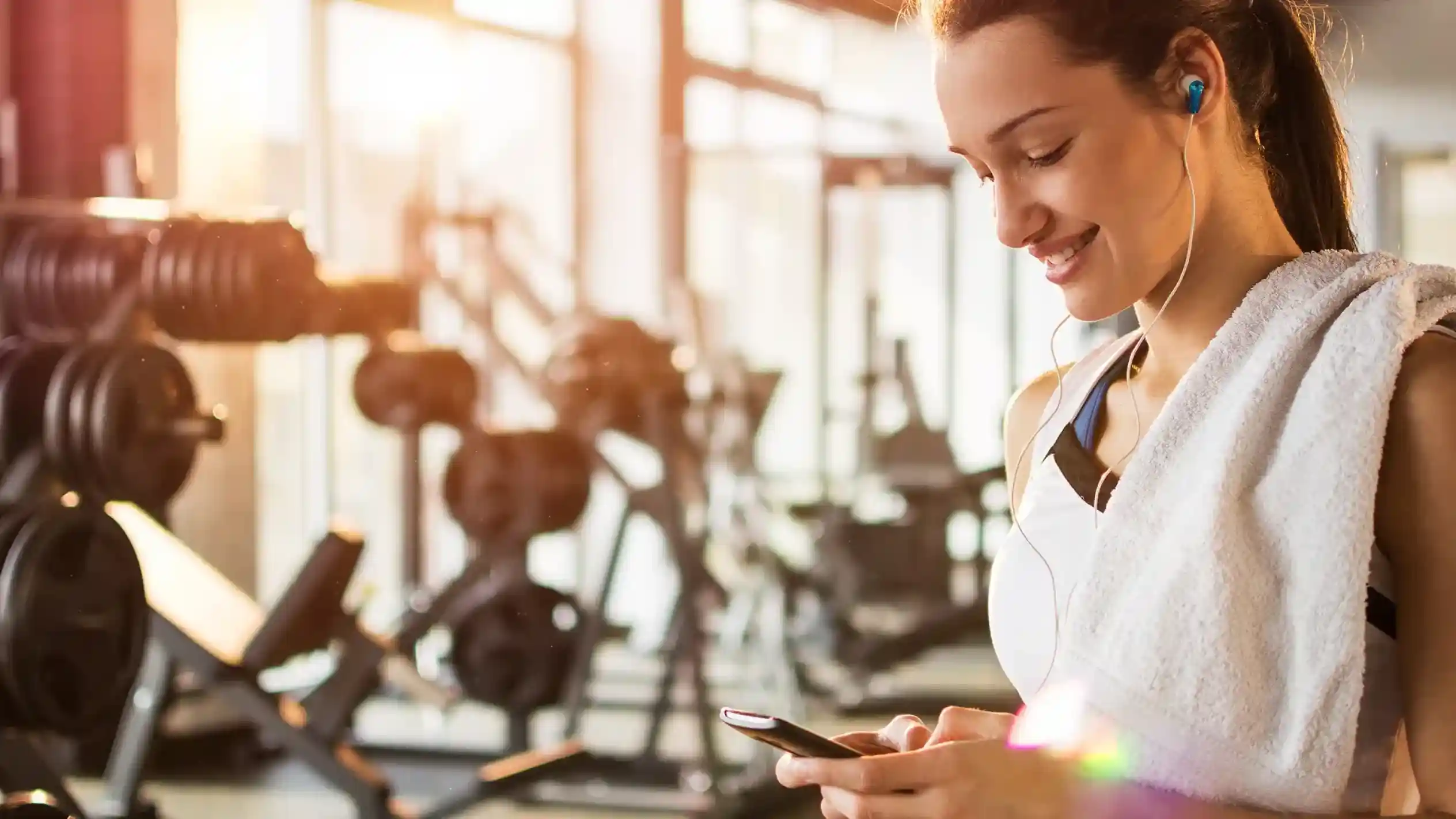 A young woman standing in a gym, wearing headphones and a workout outfit, smiles while looking at her smartphone. A towel is draped over her shoulder, and gym equipment is visible in the background.