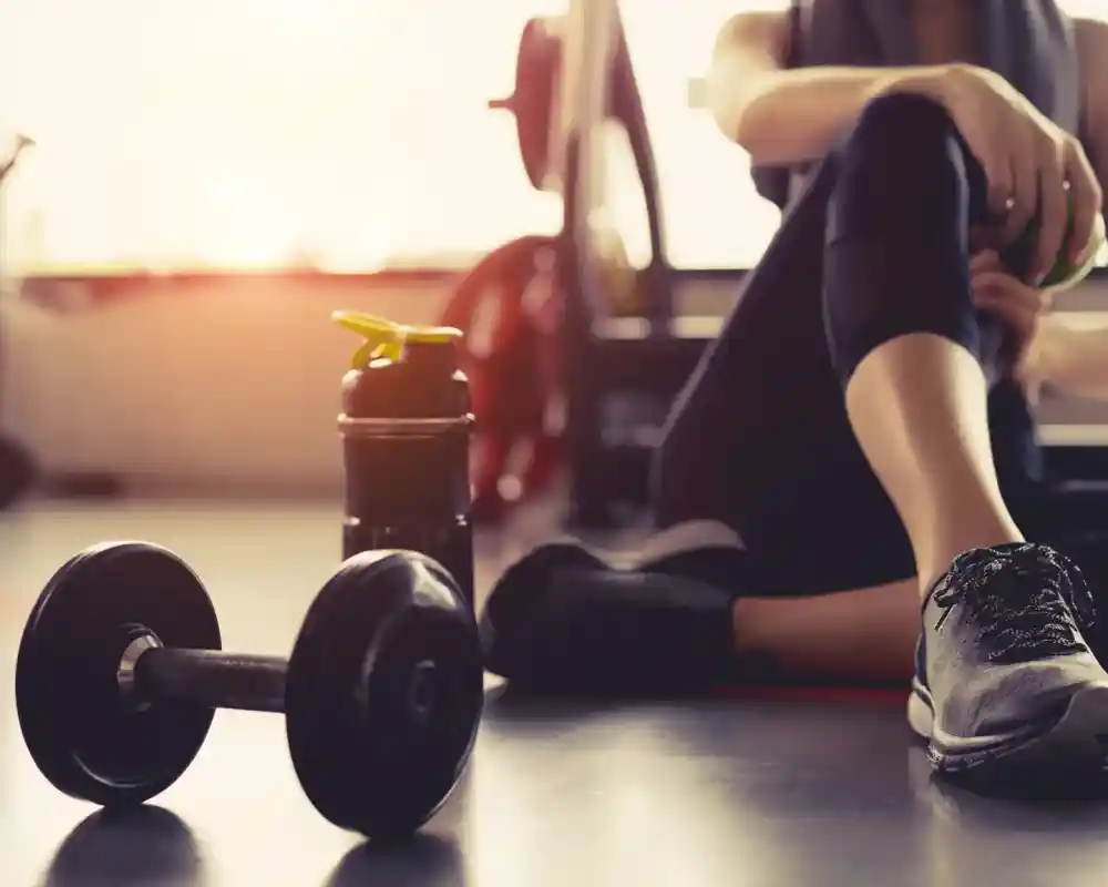 A person seated on a gym mat, resting after a workout. A dumbbell and a water bottle are placed on the floor nearby. Natural light filters in from a window, creating a warm atmosphere.
