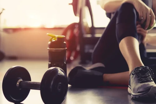 A person seated on a gym mat, resting after a workout. A dumbbell and a water bottle are placed on the floor nearby. Natural light filters in from a window, creating a warm atmosphere.