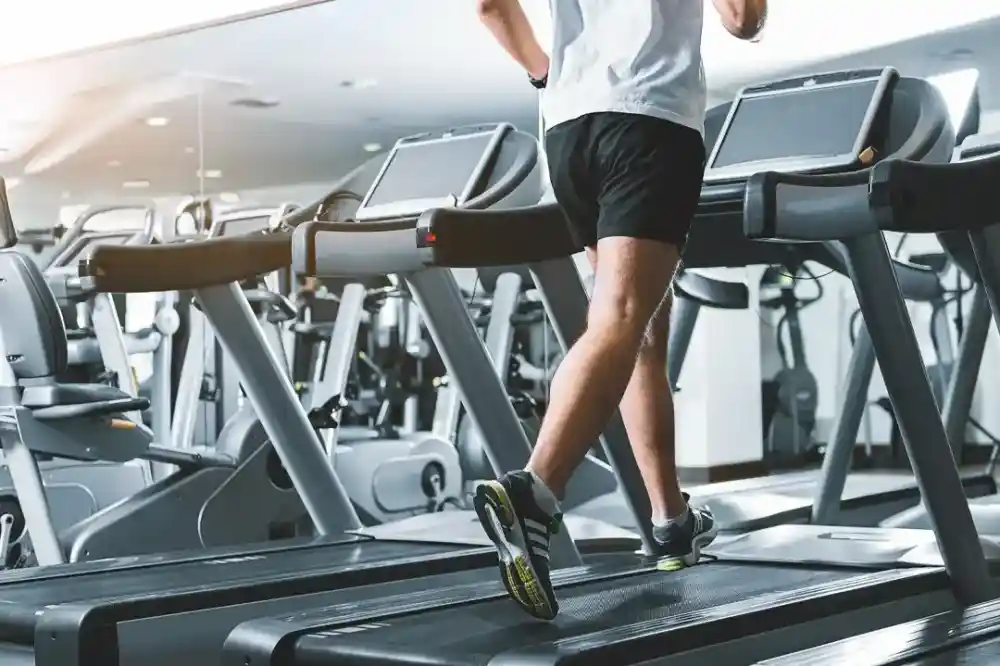 A person running on a treadmill in a gym, wearing athletic shorts and shoes, with several other treadmills visible in the background. Natural light streams into the space, reflecting off the equipment.