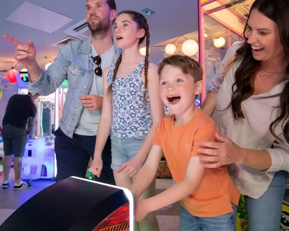 A family enjoys a fun day at an arcade, standing in front of a game. A man points excitedly while a girl looks on with wide eyes. A young boy smiles joyfully and a woman stands behind him, placing her hand on his shoulder. Colorful lights and arcade games are visible in the background.