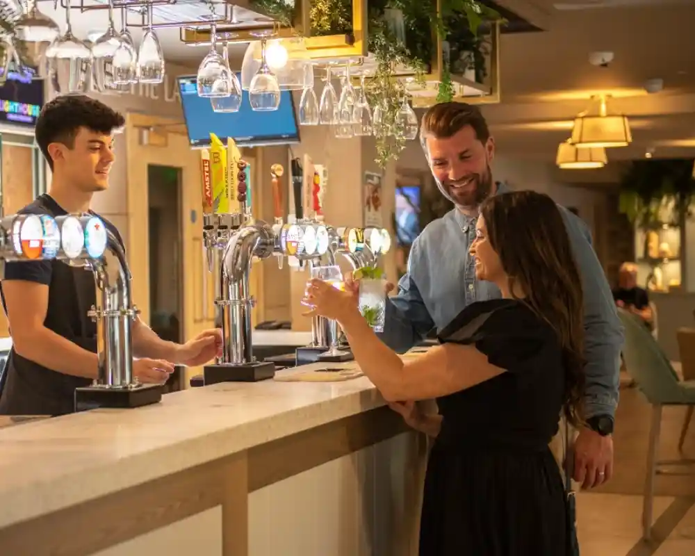 A bartender serves a drink to a woman at a bar while a man looks on, both smiling. The bar features multiple taps and a modern interior with greenery and soft lighting.
