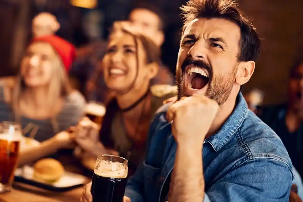 A group of people celebrating in a lively setting, with a man in the foreground passionately cheering and raising his fist. He holds a pint of dark beer, while others in the background smile and engage joyfully.