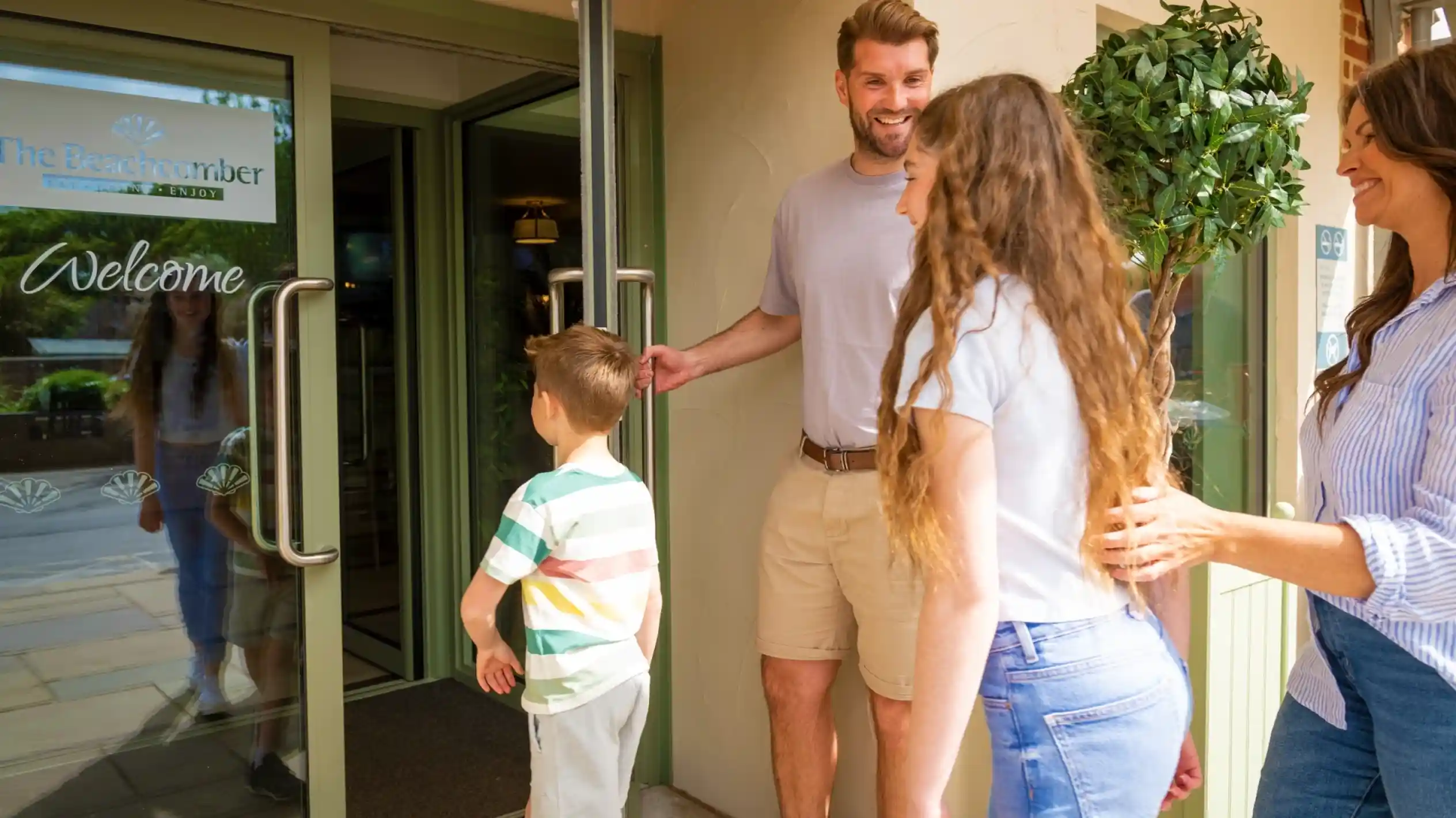 A family stands at the entrance of a welcoming resort, with a smiling man holding the door open for a woman and a young boy. The woman has long, curly hair and a friendly expression. The boy looks back at the entrance while a second woman stands beside the man, smiling.