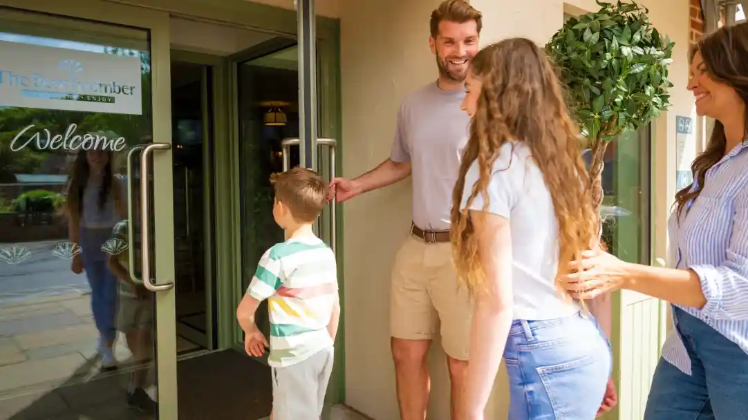 A family stands at the entrance of a welcoming resort, with a smiling man holding the door open for a woman and a young boy. The woman has long, curly hair and a friendly expression. The boy looks back at the entrance while a second woman stands beside the man, smiling.