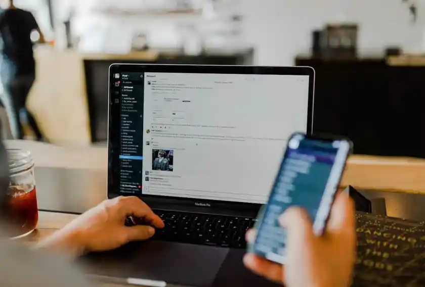 A person working on a MacBook Pro while holding a smartphone, with a Slack workspace open on the laptop screen. A glass jar with a red drink is on the left, and a book titled 