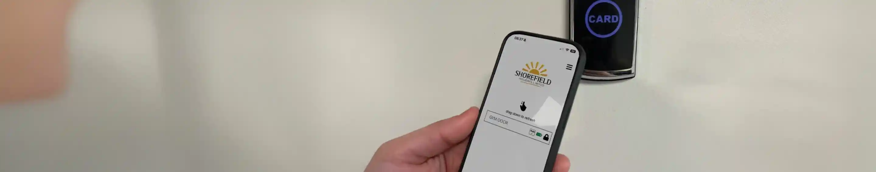 A person holding a smartphone displaying an app interface while interacting with a card reader mounted on a wall. The card reader shows a blue light indicating it's ready.