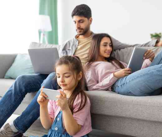 A family sitting on a couch, with a father working on a laptop, a mother using a tablet, and a young girl playing a game on her smartphone. The living room has a bright, cozy atmosphere.