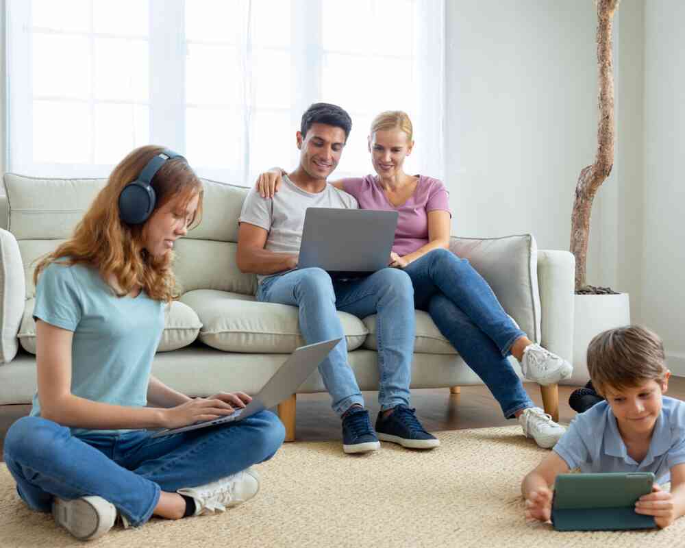 A family is sitting in a cozy living room. A woman and a man are cuddling on a sofa while using a laptop. A girl is sitting on the floor with headphones and working on her own laptop. A young boy is lying on the floor, engaged with a tablet. Natural light fills the space through the windows.