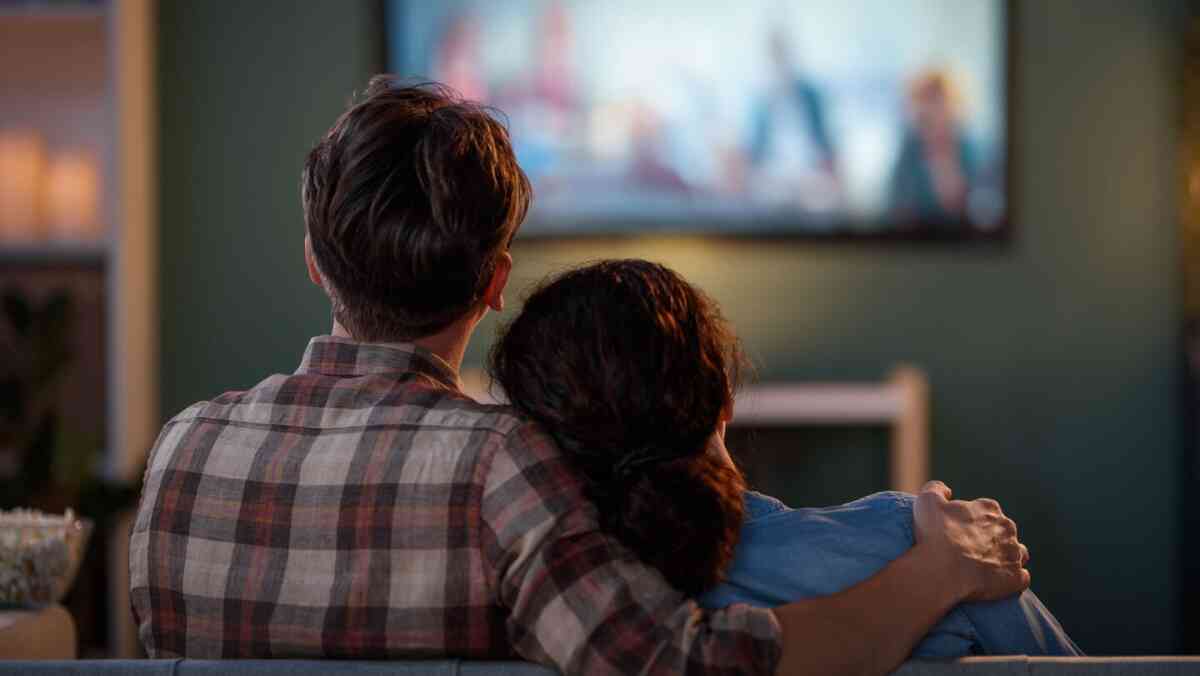 A person working on a MacBook Pro while holding a smartphone, with a Slack workspace open on the laptop screen. A glass jar with a red drink is on the left, and a book titled 