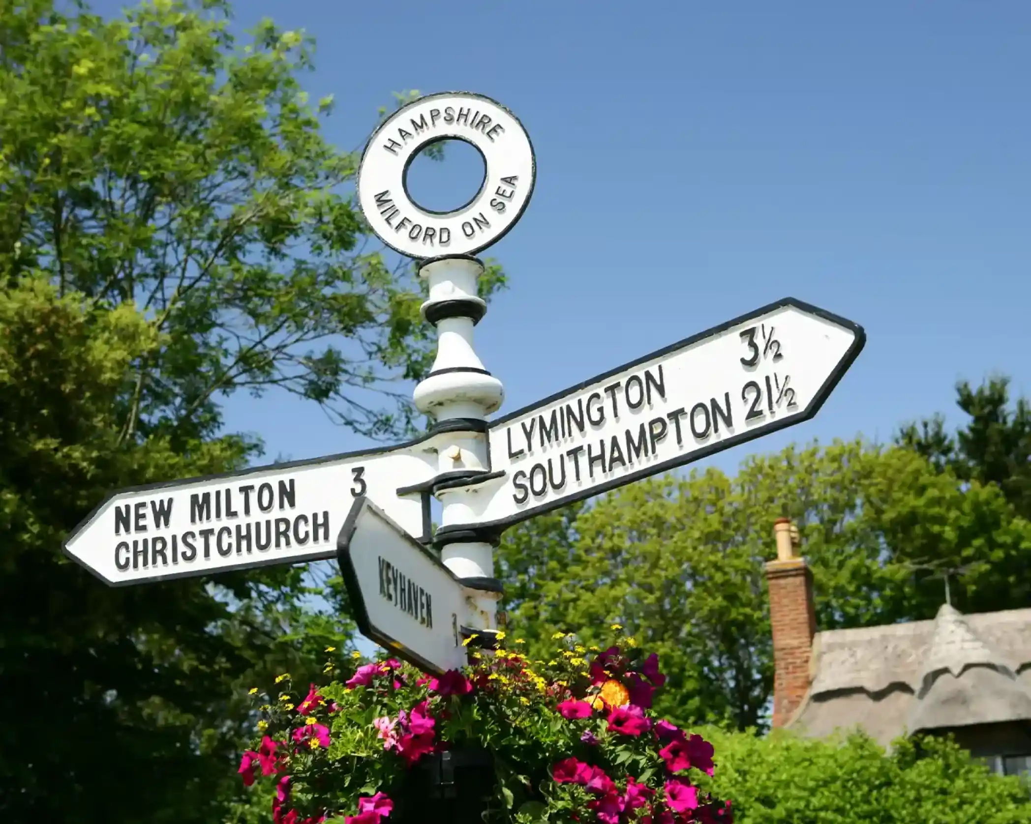 A traditional white signpost points towards Milford on Sea, New Milton, Christchurch, Lymington, Southampton, and Keyhaven, with a thatched roof cottage visible in the background.