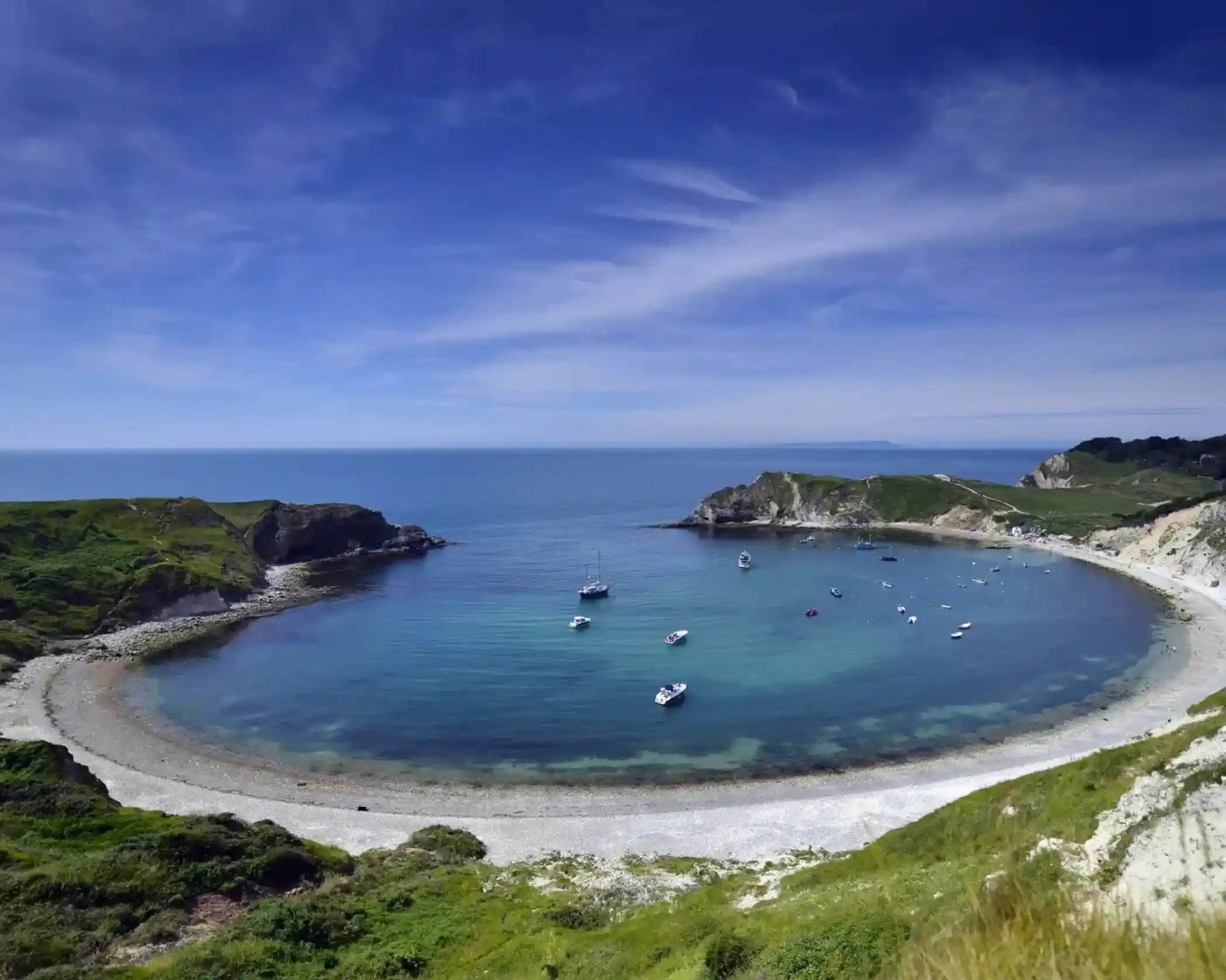 A scenic bay with several boats anchored in the clear turquoise water, framed by green, grassy cliffs under a bright blue sky.